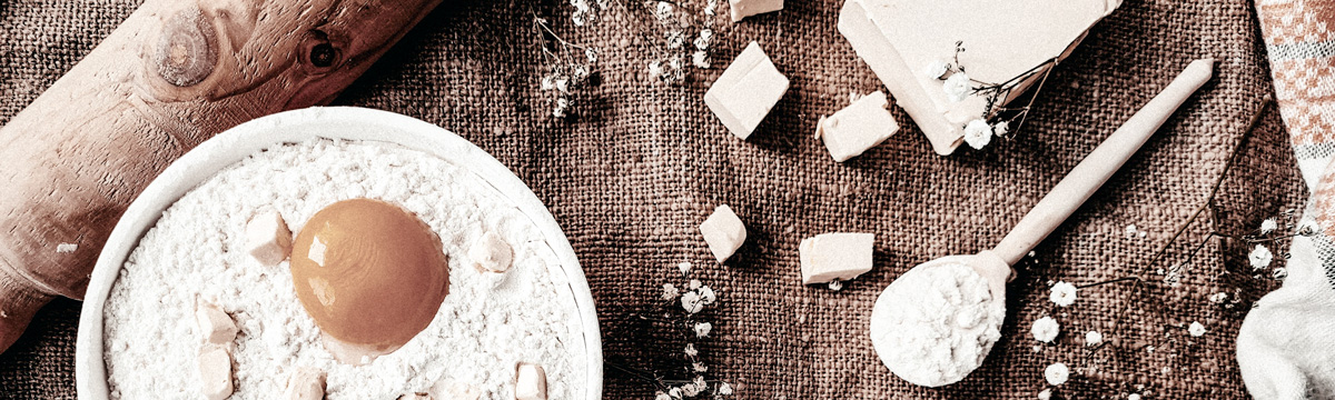 banner image: baking ingredients containing bowl 
            with raw egg in flour with pieces of lard, rolling pin, wooden spoon on a burlap background